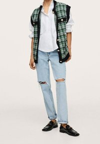 Vrouw in een witte blouse, een mouwloze vest in groen en zwart geruit met gouden knopen, gescheurde lichtblauwe spijkerbroek en zwarte Instappers.