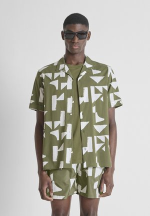 Uomo che indossa camicia e pantaloncini coordinati verde oliva con motivi geometrici bianchi, maglietta verde e occhiali da sole neri.