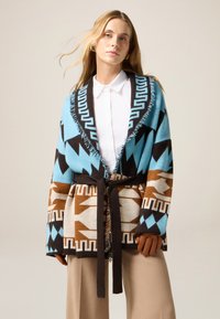 Cardigan lavorato a maglia in blu, marrone e beige con motivi geometrici. Presenta un orlo frangiato e una cintura legata in vita. Maniche lunghe e apertura frontale.