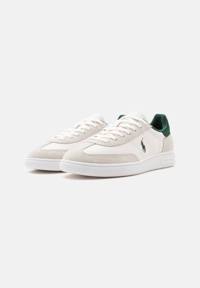 Paire de baskets basses blanches avec des accents en daim beige, une doublure intérieure verte, des lacets et un logo cheval vert sur le côté.