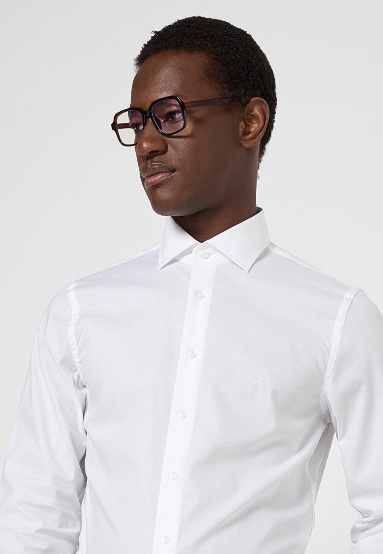 Hombre con gafas grandes de carey y una camisa blanca abotonada, mirando ligeramente de lado contra un fondo plano.