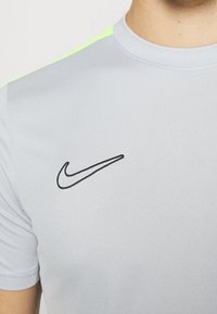 Camiseta atlética gris de manga corta, con el logo negro de Nike en el lado izquierdo del pecho y detalles en verde neón en la costura del hombro.