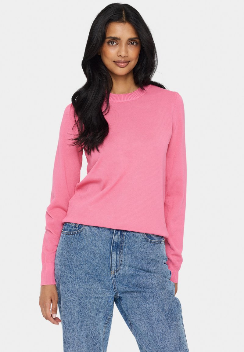 Roza pletena srajca z dolgimi rokavi in ribastim ovratnikom, v kombinaciji z modrimi trapericami visoko v pasu. Tkanina se zdi mehka z rahlo teksturo.