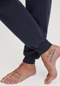 Marineblaue Fleece-Jogginghosen mit gerippten Bündchen, kombiniert mit einem barfuß, das mehrere Tattoos zeigt, darunter Text und einfache Designs.