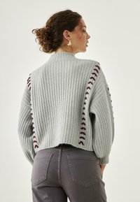 Suéter de canalé gris claro con costuras de acento burdeos a lo largo de las mangas, con un cuello alto y un dobladillo trasero recortado. Pantalones vaqueros grises.