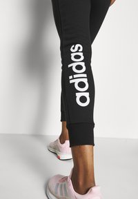 Schwarze Jogginghosen mit weißem „adidas“-Logo entlang des Beins. Kombiniert mit hellrosa Sportschuhen mit weißen Akzenten und strukturiertem Obermaterial.