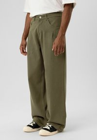 Olijfgroene, wijde broek met een gestructureerde afwerking, voorzien van voorplooien en zijzakken. Gecombineerd met zwarte en crèmekleurige sneakers.