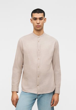 Νέος άνδρας με κοντά μαλλιά, φορώντας μπεζ μακρυμάνικο πουκάμισο με μανταρίνι γιακά και ανοιχτό μπλε τζιν, στέκεται μπροστά σε λευκό φόντο.