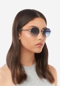 Ümarad päikeseprillid hõbedase metallraami ja sinaka gradueeritud läätsedega. Õhukesed, elegantsed sangad täiendavad minimalistlikku disaini.