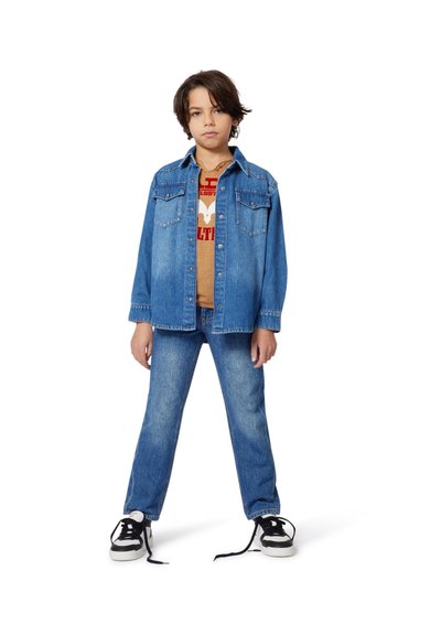 Veste en jean avec poches à rabat et un t-shirt graphique beige en dessous ; associée à un jean bleu et des baskets noires avec des semelles et des lacets blancs.