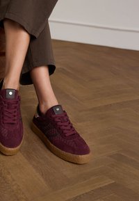 Burgunderfarbene Wildlederschuhe mit strukturierten Akzenten, schwarzen Streifen und braunen Gummisohlen, kombiniert mit kurzen, dunklen Hosen auf einem Holzboden.