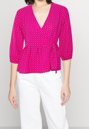 Žena nosící jasně růžovou vzorovanou zavinovací blůzu s tříčtvrtečními rukávy, doplněnou o bílé kalhoty s vysokým pasem, na jednobarevném pozadí.