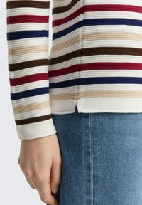 Hand naast zich rustend, gekleed in een geribde trui met horizontale strepen in crème, rood, blauw, beige en bruin, gecombineerd met blauwe spijkerbroek.