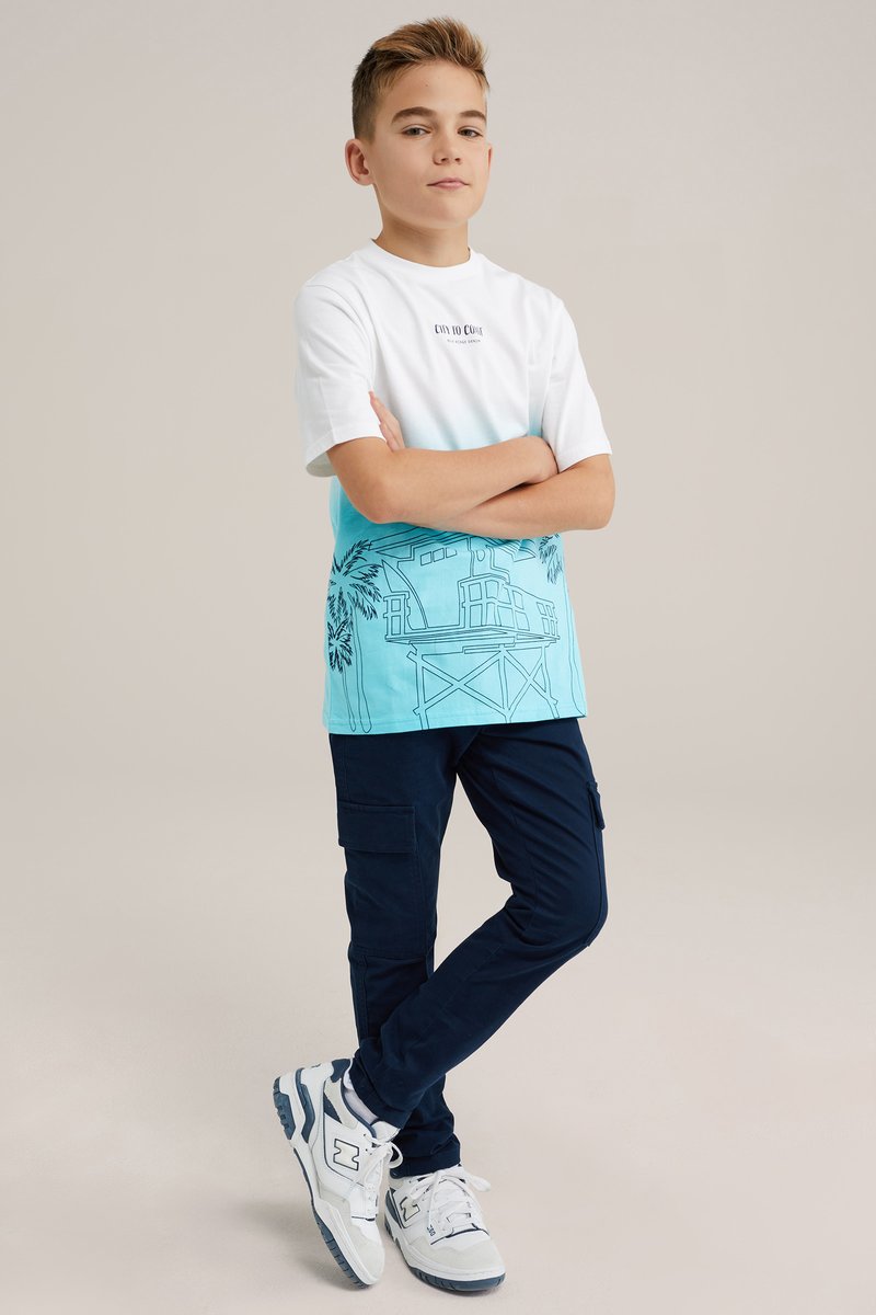 Balta un gaiši zila gradienta T-kreklu ar palmu un ēku siluetēm, apvienojot ar tumši ziliem biksēm un baltām sporta apaviem.