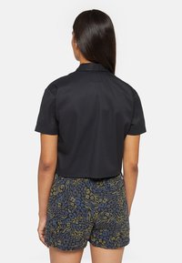 Gekürztes schwarzes Hemd mit kurzen Ärmeln und Kragen, kombiniert mit gemusterten Shorts mit grünen und blauen abstrakten Designs.
