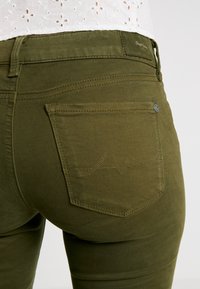 Jeans skinny verde oliva con un bolsillo trasero texturizado que presenta costuras en zigzag. La tela parece suave con un ligero brillo.