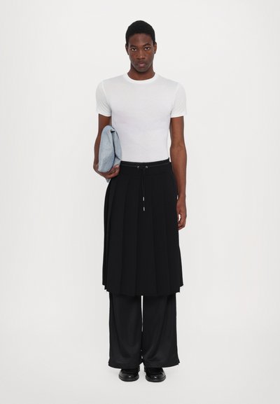 Gonna plissettata nera abbinata a una t-shirt bianca attillata, mentre tiene una borsa grigia piegata. I pantaloni a gamba larga neri e le scarpe nere completano il look.