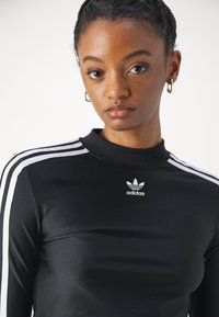 Top de manga longa preto, feito de material suave, com detalhes em três riscas brancas nos ombros e um pequeno logo branco da Adidas no peito.