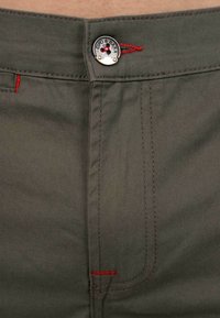 Pantalon en coton vert olive avec un bouton en métal portant le logo et des accents de surpiqûres rouges sur la taille et les coutures des poches.