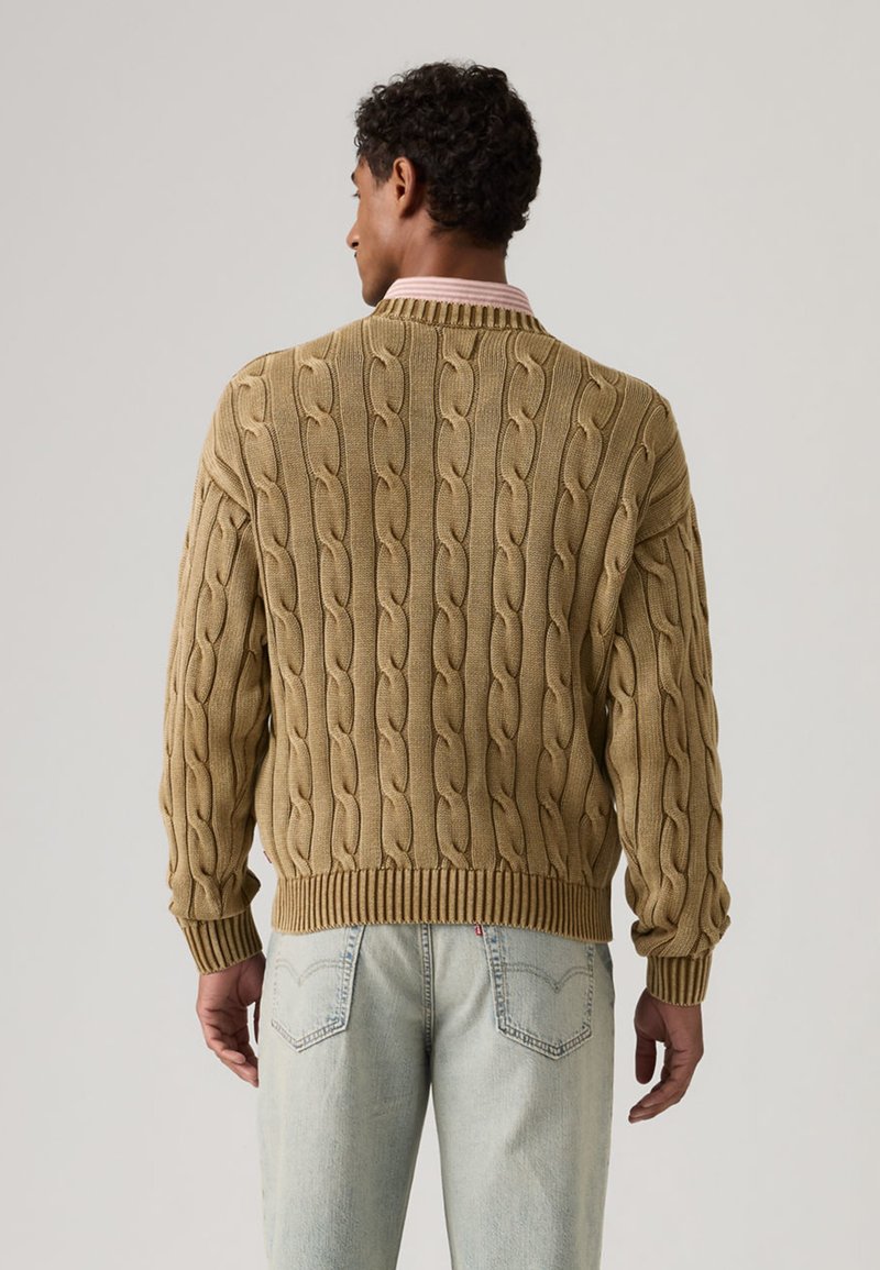 Man wearing a tan cable-knit sweater and light blue jeans, facing away against a plain light background.