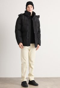 Black puffer jacket with a high collar, front buttons, and pockets, worn over a black top and cream-colored pants. Black sneakers complete the look.