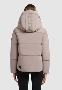 Gepolsterte beige Jacke mit großer Kapuze, die ein Logo auf der Kapuze und ein dreieckiges Etikett am Saum aufweist. Glatte Textur, eng anliegende Ärmel.