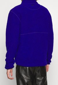Polarowy pulower w intensywnym niebieskim kolorze, z wysokim kołnierzem, prążkowanymi mankietami i kontrastowym przeszyciem. W zestawieniu z czarnymi skórzanymi spodniami.