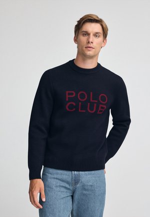Maglione a maglia blu marina con la scritta "POLO CLUB" in lettere rosse. Presenta un collo a giro e polsini e bordo a coste. Indossato con jeans in denim blu chiaro.
