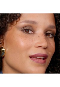 Närbild av ett ansikte med lockigt mörkt hår, varma bruna ögon, gyllene ögonskugga och en matt mauve läppfärg. Framträdande fräknar och runda guldsmycken.