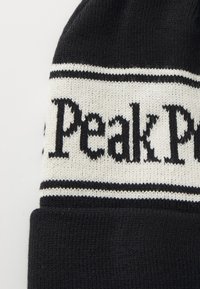 Černá pletená čepice s bílými horizontálními pruhy. Obsahuje text "Peak Performance" v tučném černém písmu na pruhu. Měkká textura.