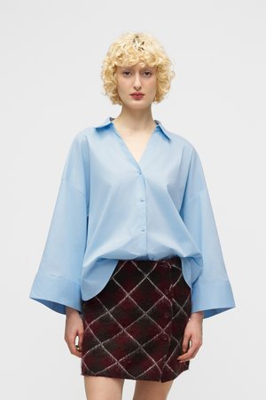 Νεαρή γυναίκα με ξανθά σγουρά μαλλιά που φοράει ένα φαρδύ γαλάζιο πουκάμισο και μια κόκκινη-μαύρη καρό μίνι φούστα μπροστά σε ένα απλό φόντο.