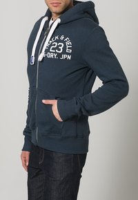 Hombre con sudadera con cremallera azul marino y cordones blancos, con el texto "Track & Field 23 Dry JPN", manos en los bolsillos delanteros, combinada con jeans oscuros.