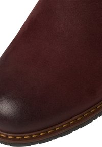 Burgundin nahkaboot, jolla on sileä pinta, pyöristetty kärki ja kontrastivärinen vaaleanruskea tikkaus yksityiskohtana pohjan ympärillä. Kuivasti teksturoitu pinta.