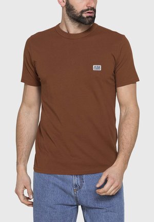 Uomo che indossa una t-shirt marrone a maniche corte con un piccolo logo sul petto e jeans di denim azzurro chiaro, guarda di lato.