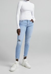 Jean skinny bleu clair avec détails usés, associé à un col roulé blanc côtelé ajusté et des baskets blanches. Texture lisse, style décontracté.