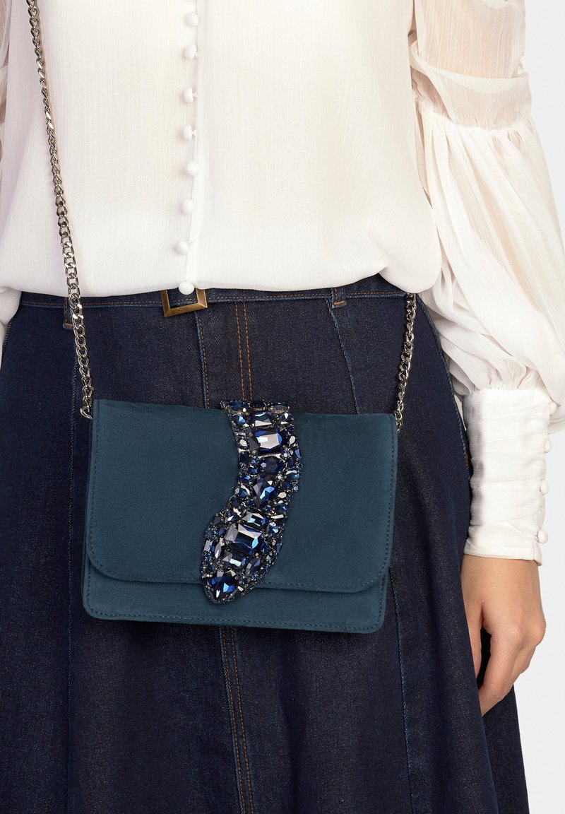 Sac en daim couleur sarcelle avec un accent décoratif en pierre précieuse bleue. Il possède un rabat plié et une bandoulière en chaîne argentée, porté avec un chemisier blanc et une jupe en jean.