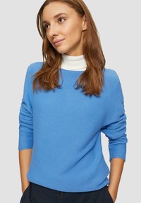 Blauwe gebreide trui met een gestructureerd design, wijde halslijn en lange mouwen, gedragen over een witte turtleneck. Geen zichtbare patronen of accenten.