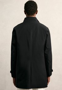Fekete, térdilő kabát sima, matt felülettel. Álló gallérral és gombos mandzsettával rendelkezik. Egyszerű, klasszikus dizájn, testre szabott szabással.