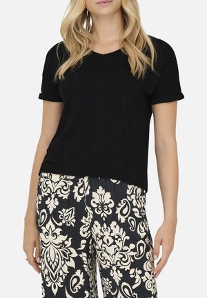 Femme portant un haut noir à manches courtes et un pantalon large à motifs noir et blanc, debout devant un fond uni.