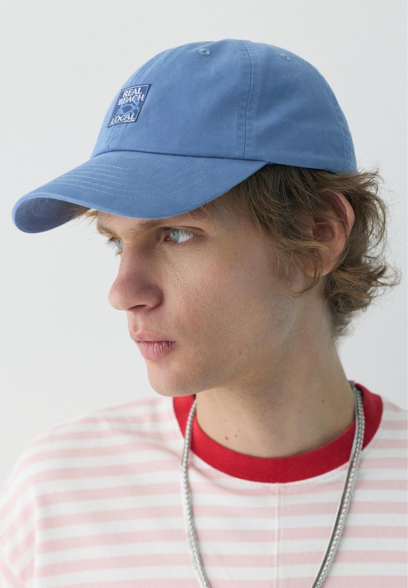 Jeune homme portant une casquette bleue avec un écusson "Real Beach Local", un t-shirt rayé rouge et blanc et une chaîne argentée, regardant sur le côté sur un fond uni.