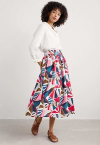 Jupe maxi multicolore avec des motifs géométriques en rose, bleu, marron et blanc ; taille froncée ; associée à un chemisier en lin blanc et des sandales marron.