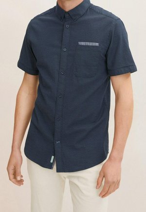 Homme portant une chemise bleu marine à manches courtes à boutons avec un motif légèrement à pois et un pantalon de couleur claire.