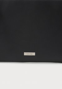 Melna ādas rokassoma ar gludu tekstūru un metāla Calvin Klein logo plāksni. Vienkāršs, taisnstūra dizains bez papildu rakstiem.