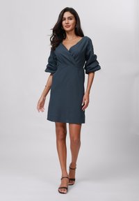 Robe vert foncé avec un décolleté en V, des manches bouffantes et un design ajusté. Texture lisse, longueur genou, accompagnée de sandales à brides de cheville noires.