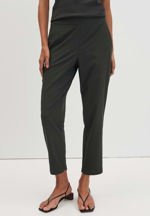 Vrouw draagt zwarte slim-fit enkelhoge broek en zwarte sandalen met bandjes en lage hak, staand tegen een effen achtergrond.