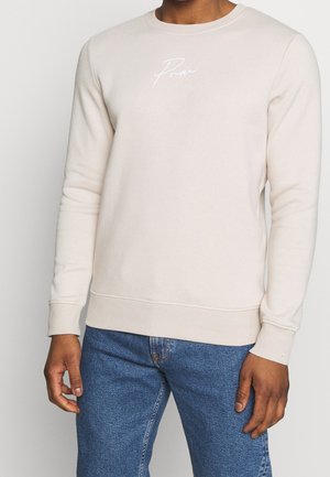 Man draagt licht beige sweatshirt met kleine witte tekst op de borst, gecombineerd met blauwe spijkerbroek, staand tegen een effen achtergrond.
