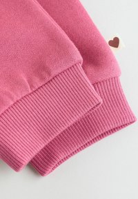 Mangas de suéter de punto rosa con puños acanalados y una pequeña etiqueta blanca con un corazón marrón sobre un fondo blanco.