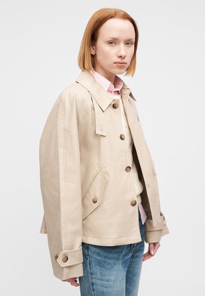 Μπεζ βαμβακερό σακάκι με κλασικό γιακά, κούμπωμα με κουμπιά και δύο τσέπες. Διαθέτει φαρδιά μανίκια και μανσέτες με κουμπιά. Φοριέται πάνω από ένα ανοιχτόχρωμο πουκάμισο.