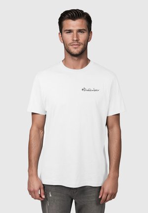 Mężczyzna o krótkich ciemnych włosach, ubrany w gładki biały T-shirt z czarnym napisem „#Problemlöser” na lewej piersi, stojący na szarym tle.