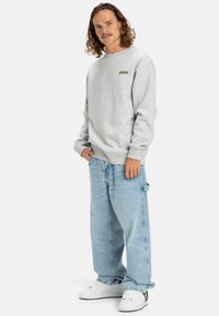 Felpa grigia con polsini a costine, caratterizzata da un piccolo logo verde. Abbinata a jeans azzurri larghi e sneakers bianche.